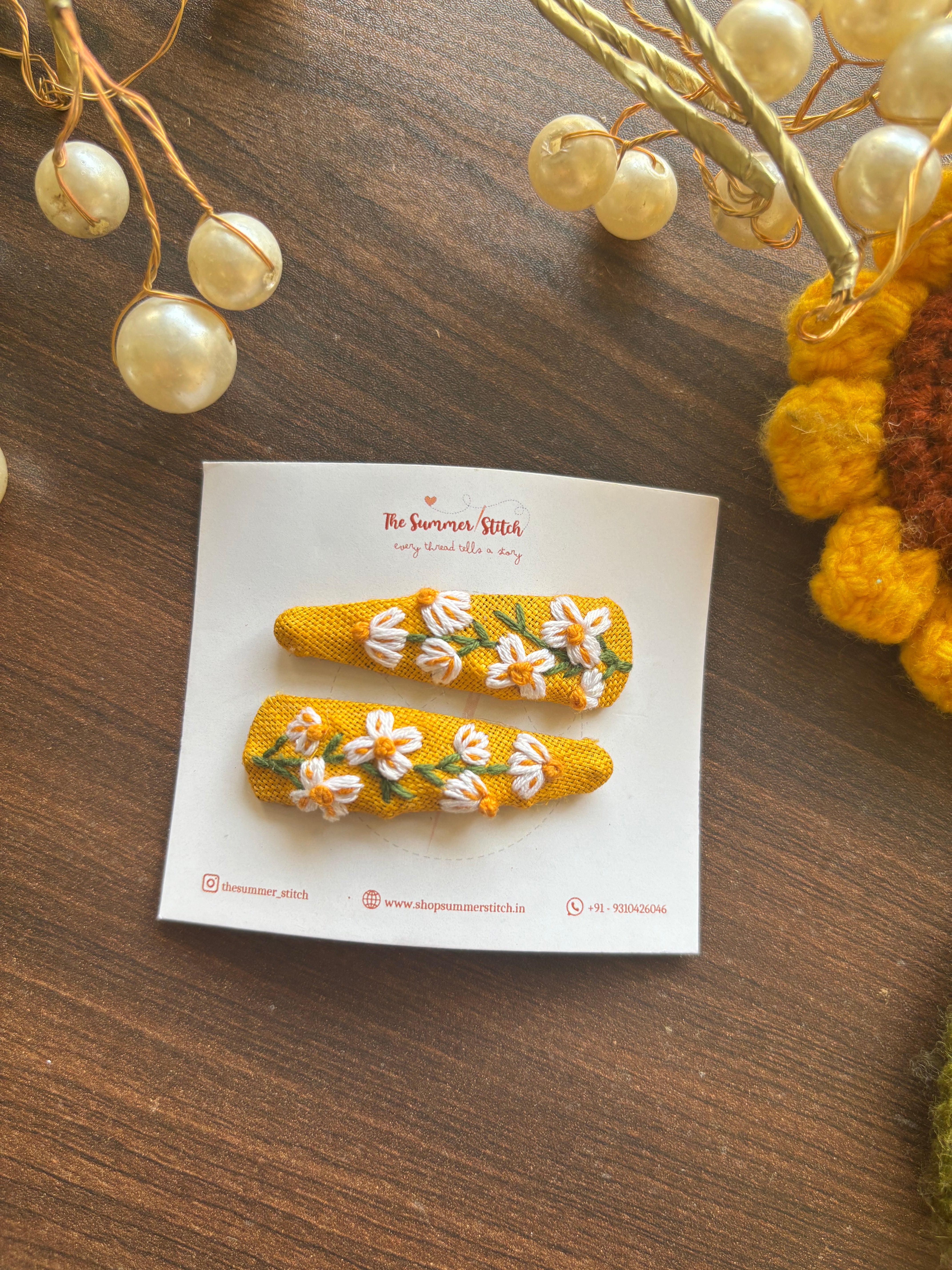 A pair of embroidered hair pins with a daisy pattern, displayed on a white card with decorative elements in the background.